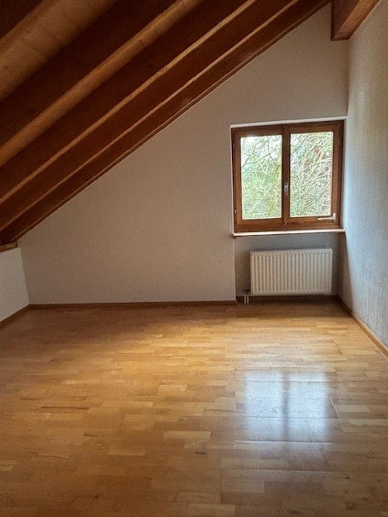 Empty attic room with wooden floors, inclined ceiling, large window, white walls, and white radiator
