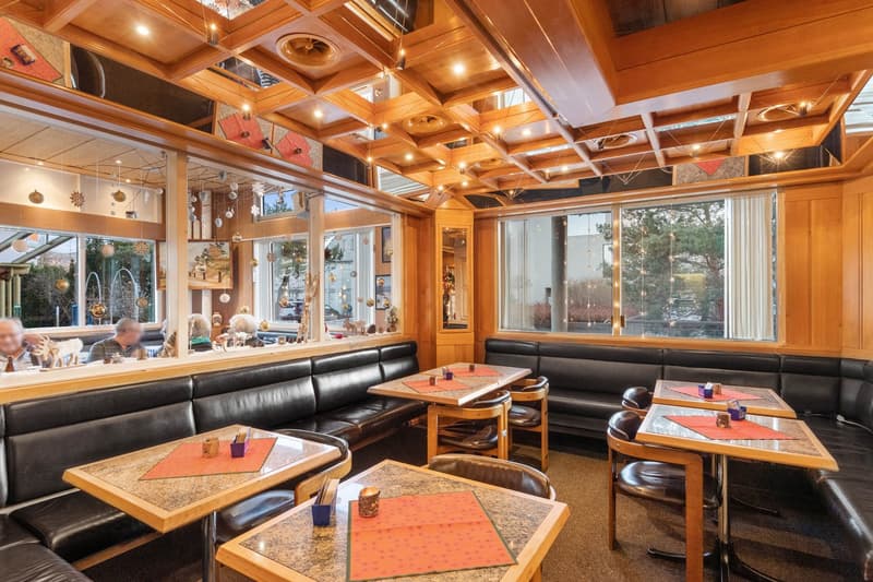 Cozy and rustic interior with wooden ceiling and walls, large windows overlooking the outdoors, leather sofas, and wooden tables and chairs. The space has a warm and inviting atmosphere, with decorative elements like hanging ornaments and a fireplace visi