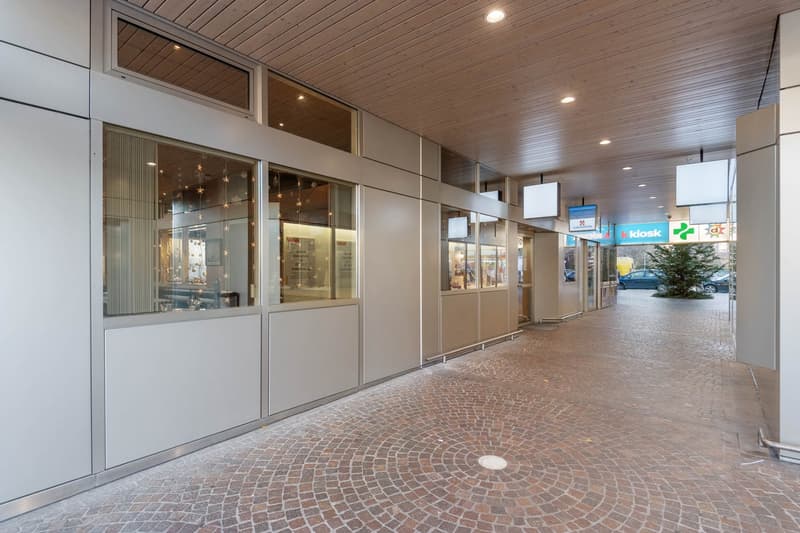 The image shows the exterior of a commercial building with a covered walkway and paved area in front. The building has large glass windows and doors, with a wooden ceiling and recessed lighting. There are various retail stores and businesses visible, incl