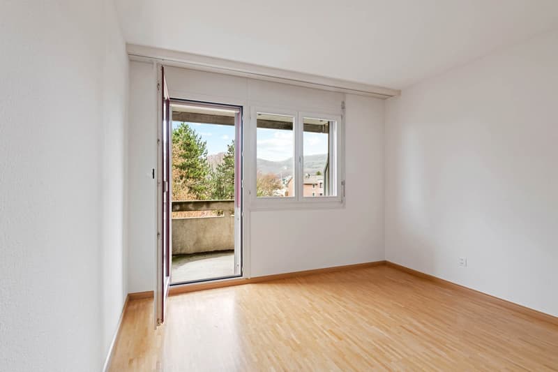 Empty room, white walls, wooden floors, glass door leading to balcony, power outlet