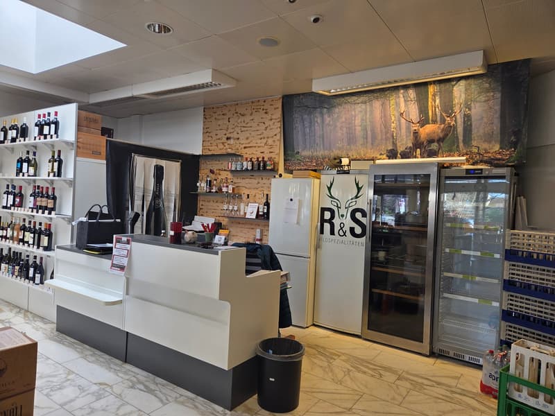 Open interior with wooden walls, tiled flooring, a reception desk, refrigerators with the text R&S, wine bottles on shelves, boxes, and a trash bin.