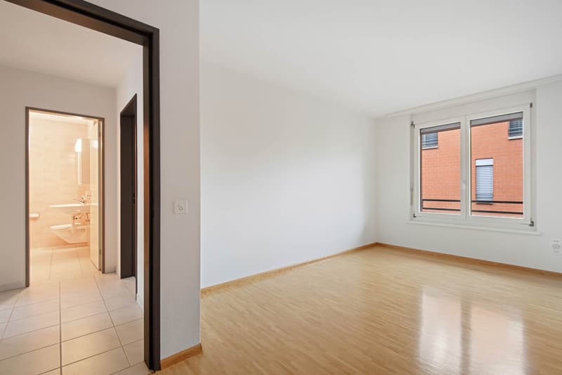 Empty room with white walls, wooden floor, and a window overlooking a brick building. The room has a door leading to a bathroom with a toilet and sink.