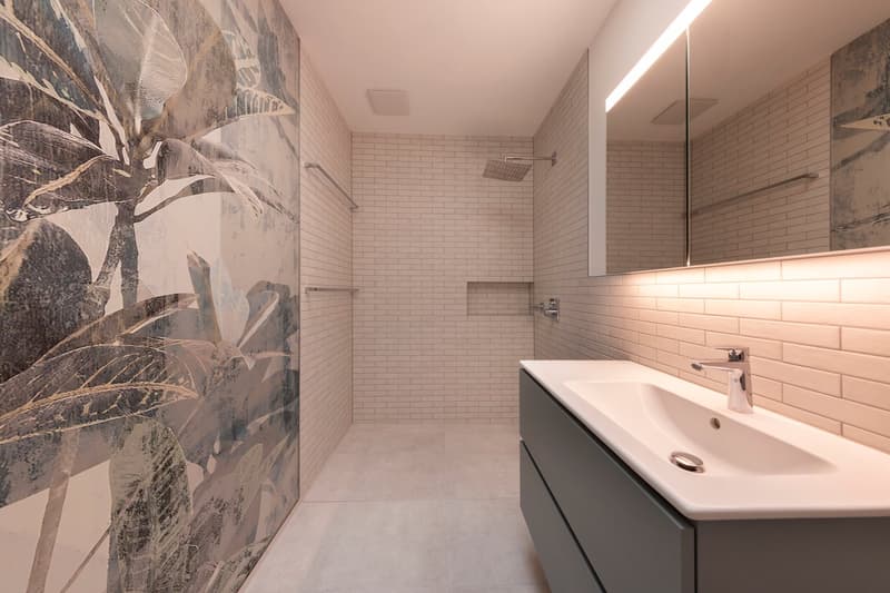 Modern shower area with large shower head, wall-mounted shower rack, gray vanity, large mirror, tiled walls, and art mural of leaves.