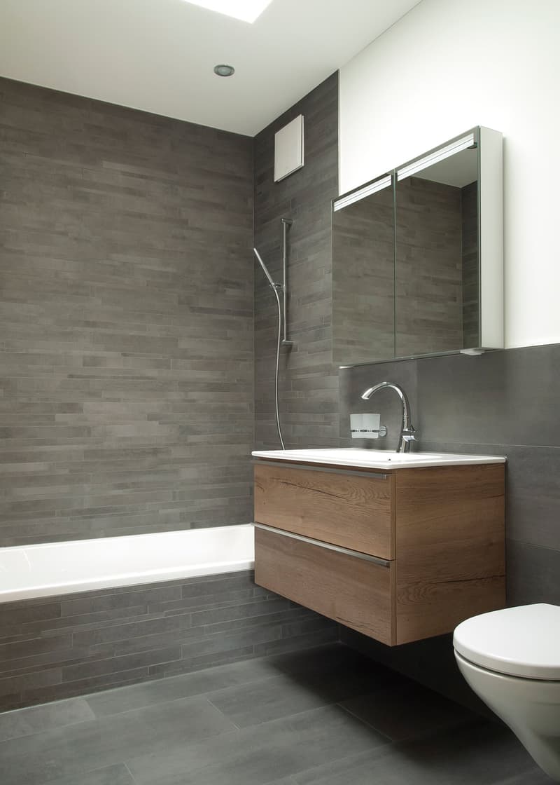 Modern bathroom with gray tile walls, wooden vanity, white bathtub, glass shower enclosure, and white ceramic sink.
