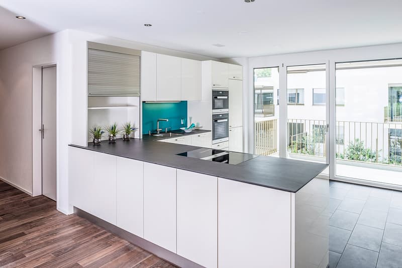 Modern open plan kitchen with island, white cabinets, black countertop, turquoise backsplash, glass door leading to balcony, built-in oven and microwave, stainless steel sink with faucet, ceramic floor.