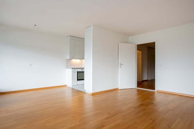 Large empty room, wooden floor, open kitchen with an oven, doorways leading to other rooms