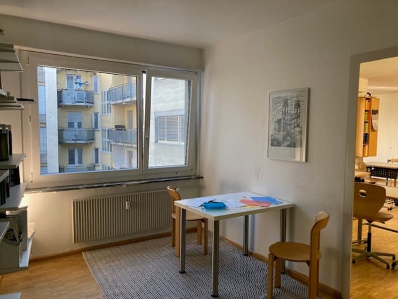 small room, wooden desk, 2 chairs, white walls, window, radiator, carpet, shelves, table, books, picture frame