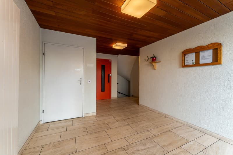 Empty hallway with white door, red door, wooden ceiling, tiled floor, wall mounted light, bulletin board