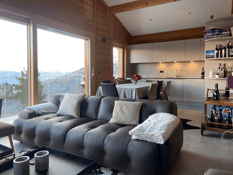 Open-concept living room and kitchen with large windows offering a scenic view, wooden walls and ceiling, gray sofa, dining table, and kitchen with cabinets and appliances