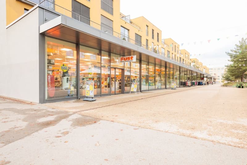 Multi-story commercial building with a large retail store called Fust occupying the ground floor. The building has a modern, glass-fronted design with balconies on the upper floors. There is a paved area in front of the building that appears to be used fo