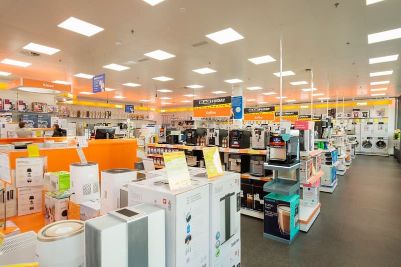 Large retail store with various home appliances and electronics on display, including coffee makers, air purifiers, and dishwashers. The store has bright lighting, orange and yellow accents, and signage for Black Friday sales.