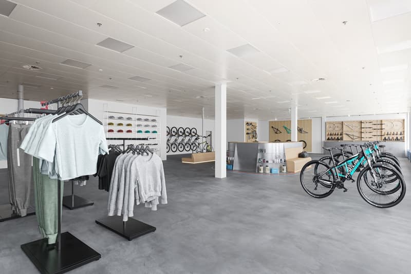 Large open space with clothing racks, shelves displaying accessories, and bicycles on display. The space has a modern, minimalist design with white walls, gray flooring, and recessed lighting.