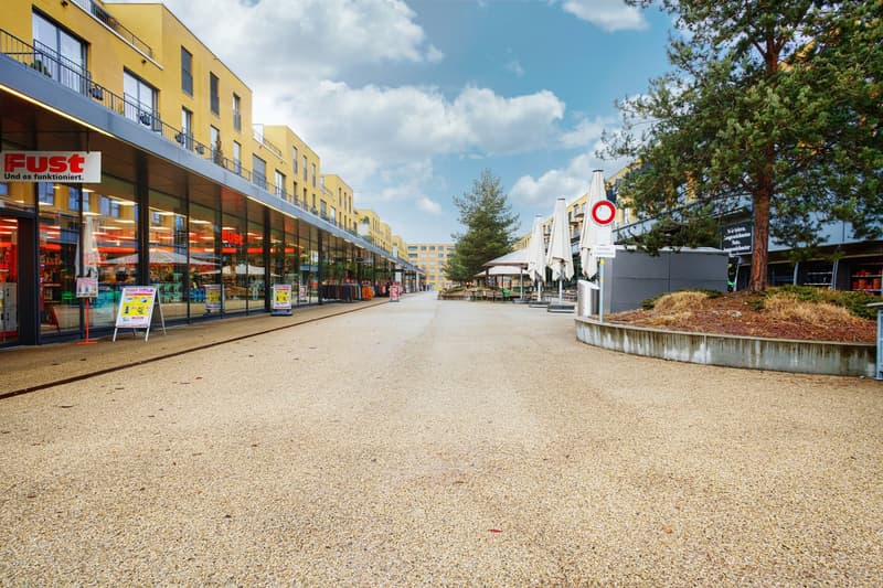 Outdoor shopping area with yellow buildings, stores, and a paved walkway with a fountain and trees