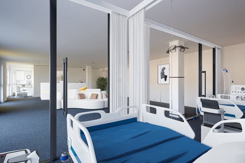 The image shows a modern and spacious hospital room with a comfortable-looking bed, white curtains, and various medical equipment such as an IV stand and a monitor. The room has large windows that provide natural light and a view, suggesting the presence