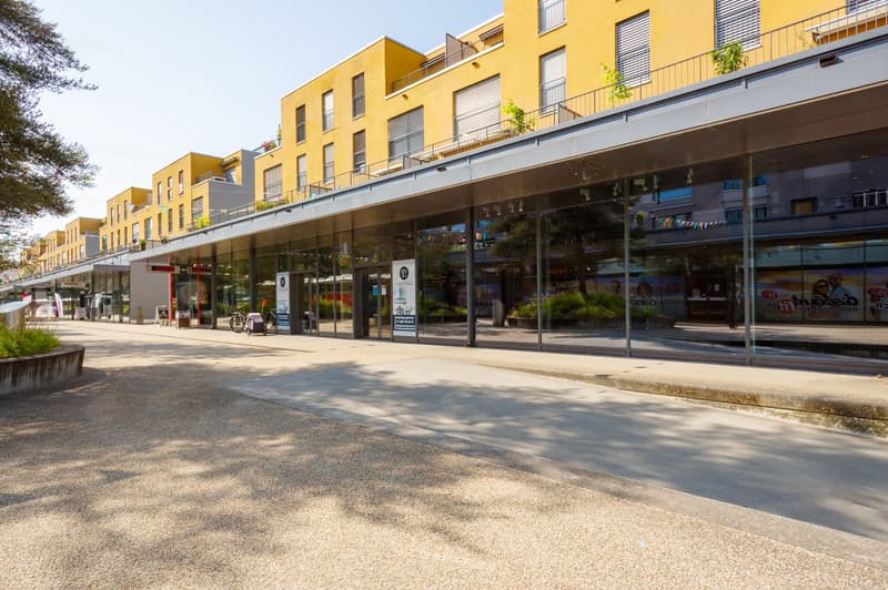 Multi-story commercial building with yellow exterior, glass storefronts, and balconies on the upper floors. There is a paved walkway in front of the building with some benches and landscaping.