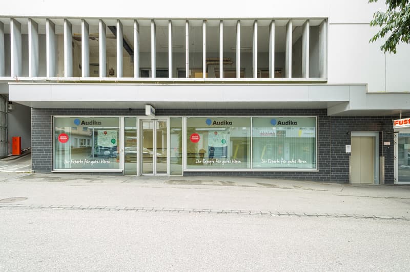 The Audika building is a white, modern structure with large glass windows, a brick wall, and white shutters.