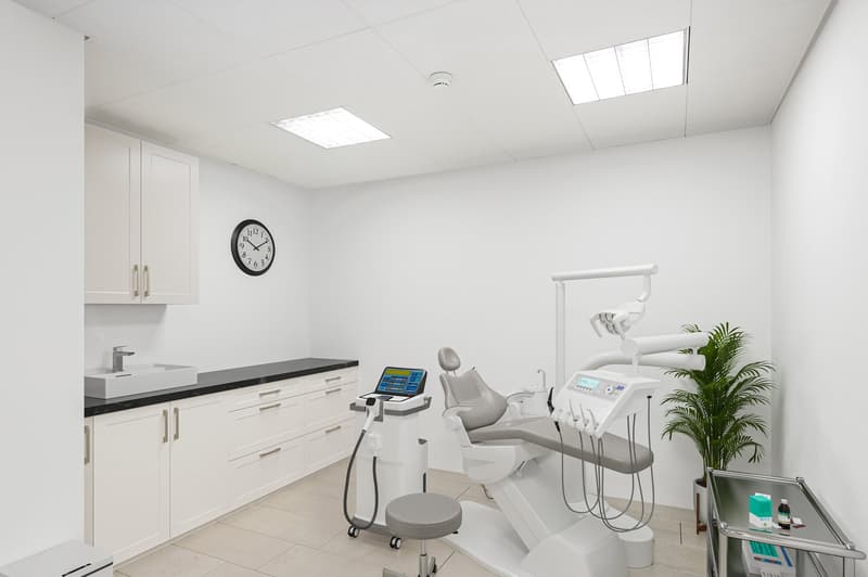 Dental clinic room with white walls, tiled floor, dental chair, sink, cabinets, clock, and a potted plant.