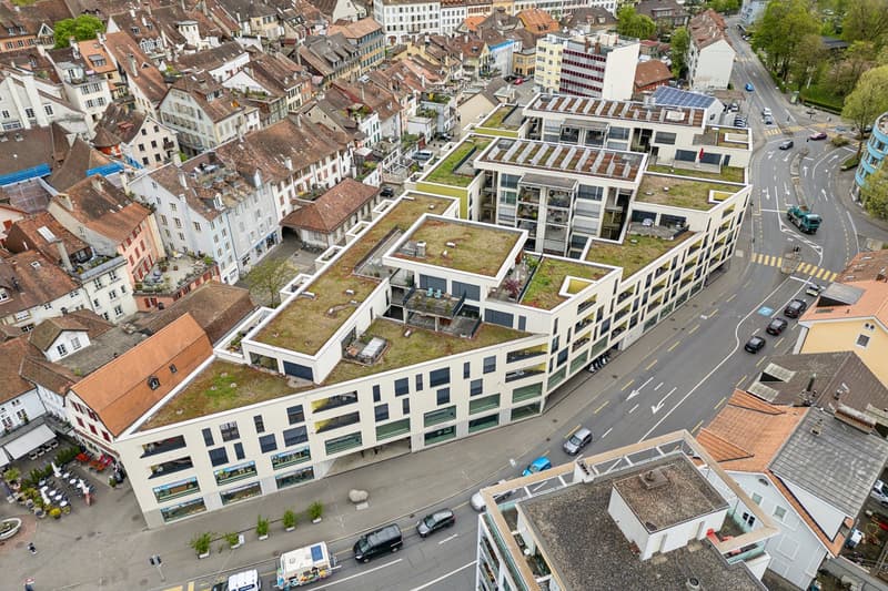 Modern residential building with green roofs, several floors, adjacent to a road with cars, surrounded by older buildings with tiled roofs