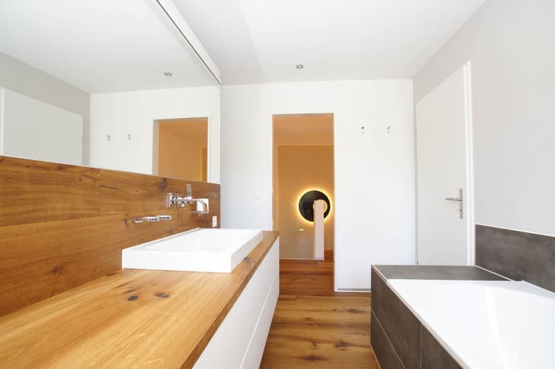 Spacious bathroom with wooden vanity countertop, white sink, and recessed lighting. Doorway leading to another room is visible.