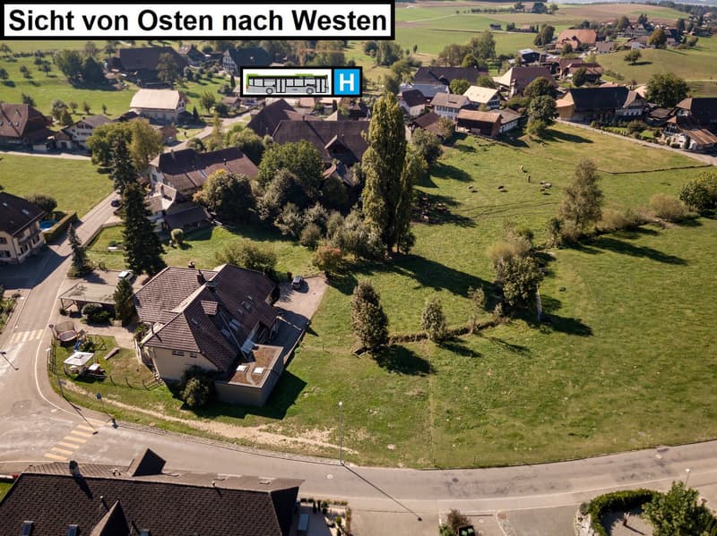 Aerial view of a village with traditional houses, green fields, and trees. There is a bus stop and some parking spaces visible in the image.