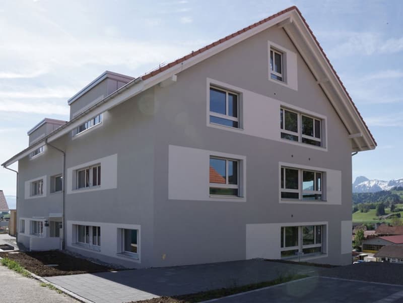 3-story apartment building with gray exterior, large windows, and a balcony visible on the upper floor