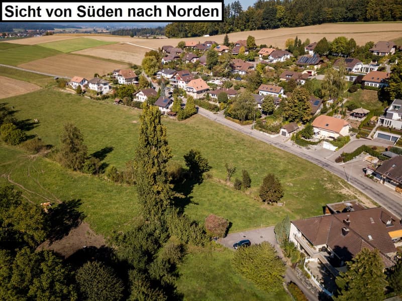 The image shows an aerial view of a small town or village in a rural area. The town is surrounded by fields and forests, with a winding road running through the center. The houses in the town appear to be a mix of traditional and modern styles, with some