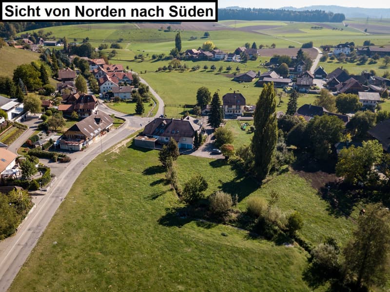 This image shows an aerial view of a rural village or town in a hilly, forested area. The landscape consists of rolling green fields, scattered houses and buildings, and trees. The town appears to have a mix of traditional and modern architecture, with so