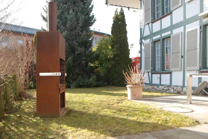 A large metal grill structure, a large plant pot with dried branches, a red telephone booth, shrubs, a fence, and a white house in the background
