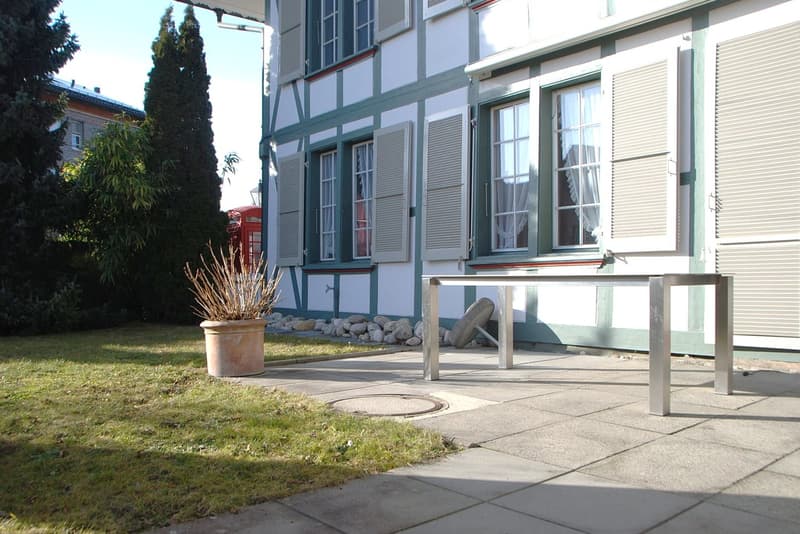 Exterior: Building with white and green painted walls, white shutters, a few windows, a table on the paved patio, and a potted plant.