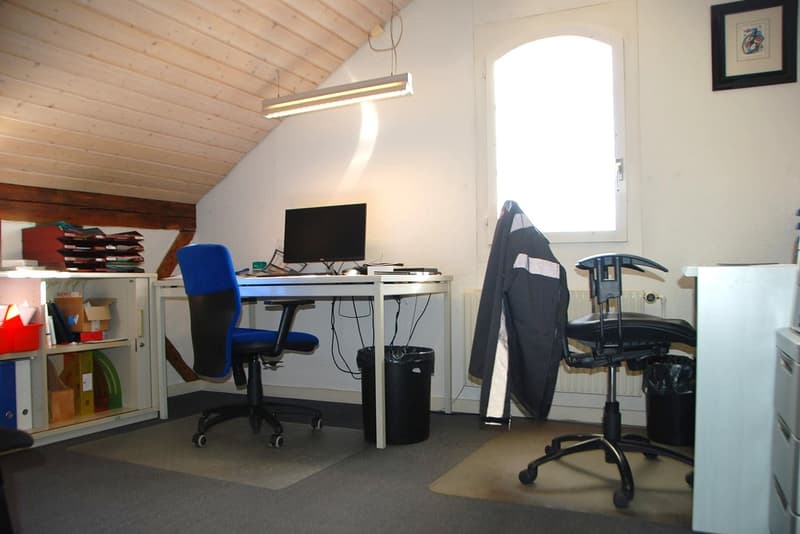 An attic office with a desk, computer monitor, and chairs. A wooden shelf on the left has office supplies. The walls are white, and there is a window on the right with a picture frame.
