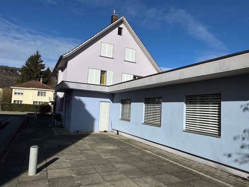 Two-story house, light blue exterior, white shutters, raised ground floor, concrete walkway