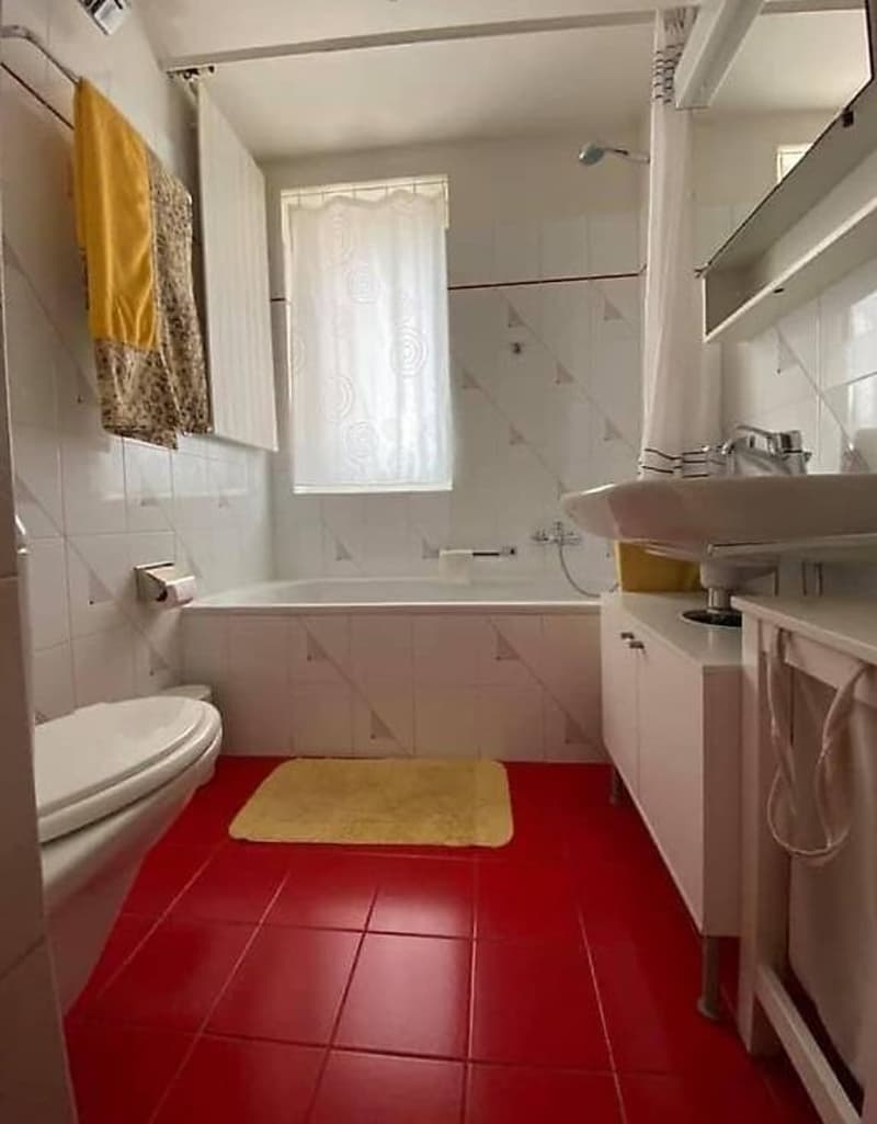 Bathroom with white tiled walls, red tile floor, bathtub, toilet, vanity with sink, and a washing machine visible.