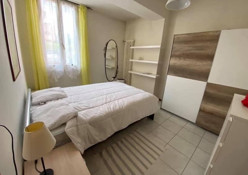 Spacious bedroom with a large bed, white bedding, and a wooden wardrobe. The room has large windows with yellow curtains, providing natural light. There is also a mirror and shelving unit in the room.