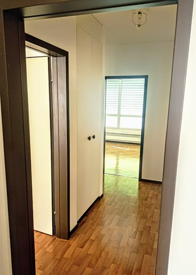 Hallway with hardwood floors, white walls, and a window with blinds. There are two doors leading to other rooms.