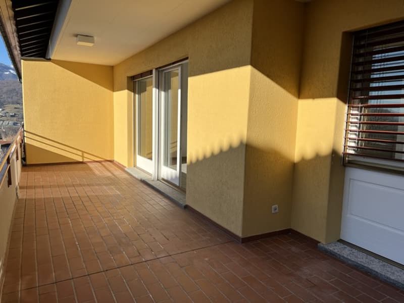 The image shows a spacious balcony or terrace with a tiled floor and sliding glass doors leading to the interior. The walls are painted in a warm, yellow-toned color, and the space appears to be well-lit with natural light.