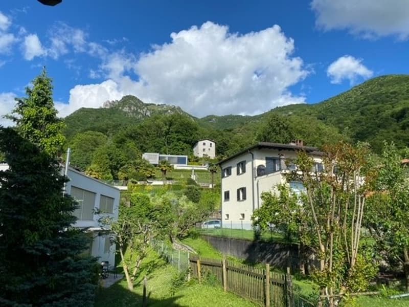 The image shows a scenic mountain landscape with lush green hills and trees. In the foreground, there are several buildings, including a white house and other residential structures, nestled among the greenery. The sky is blue with fluffy white clouds, cr