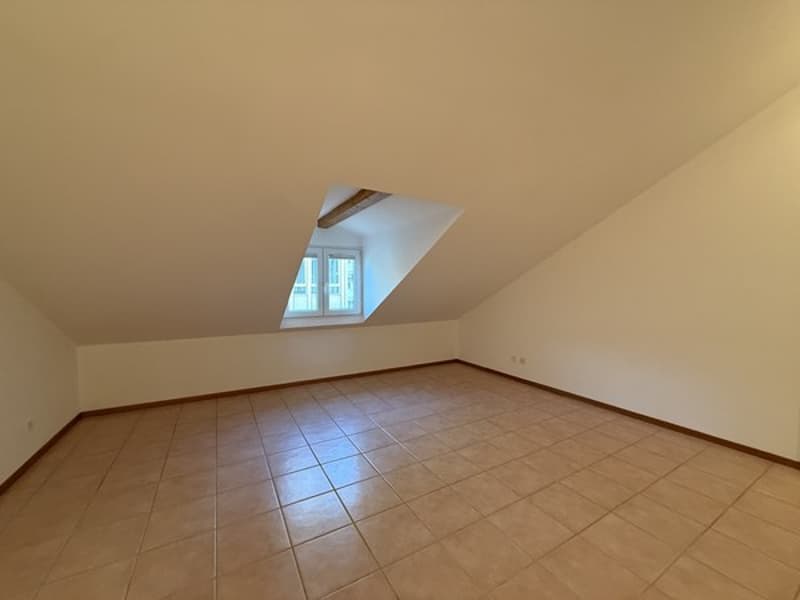Empty room with tiled floor, white walls, ceiling beams, and a small attic window