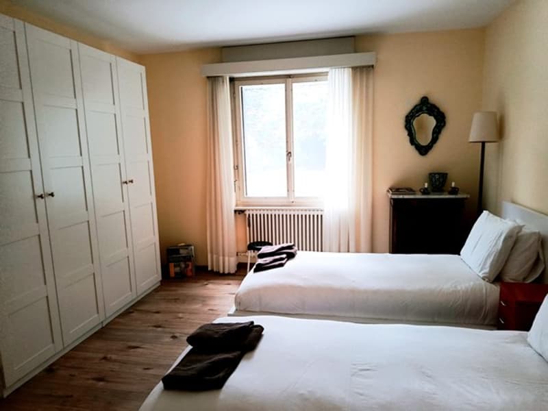 bedroom with two single beds, wooden floor, large wardrobe, mirror, radiator, window with white curtains, nightstands, table lamp, side table