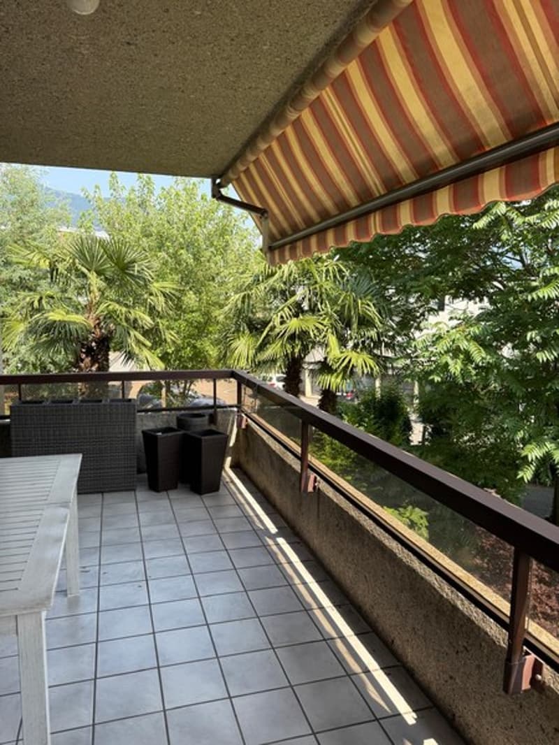 Covered balcony with striped awning, overlooking a lush garden with palm trees