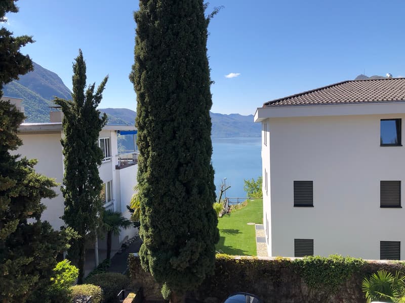 Two white buildings, one with a balcony, surrounded by greenery, with a lake and mountains in the background.