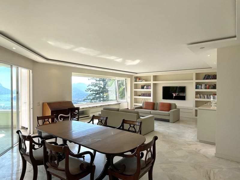 Spacious room with a dining table, chairs, couch, ottoman, bookshelf, TV, and a glass on the counter. Large windows offer a scenic view.