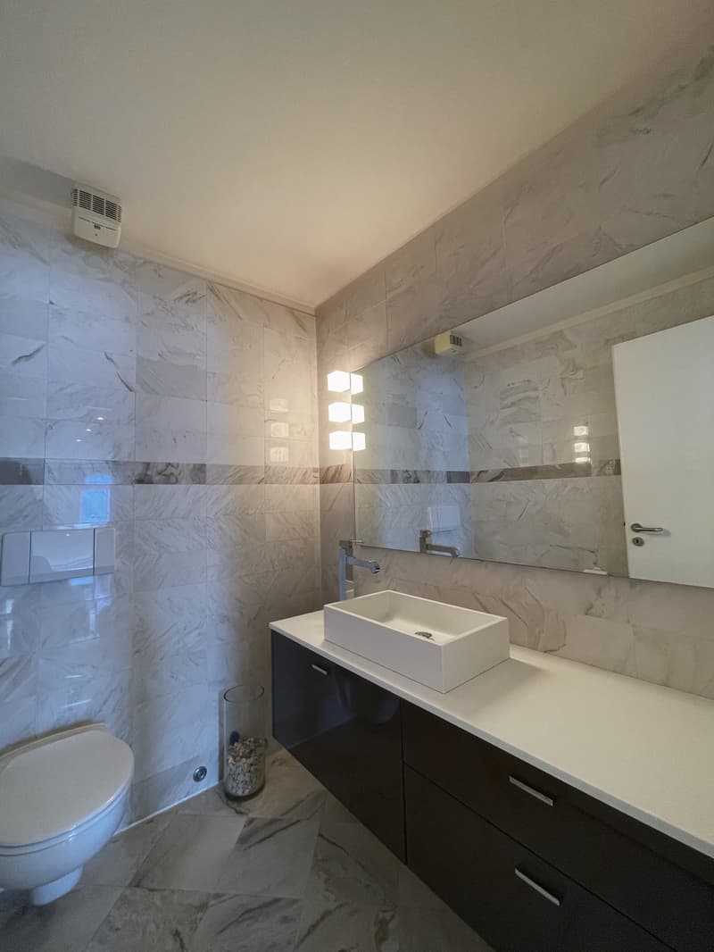 Modern bathroom with marble tiles, white sink, large mirror, and dark cabinets.