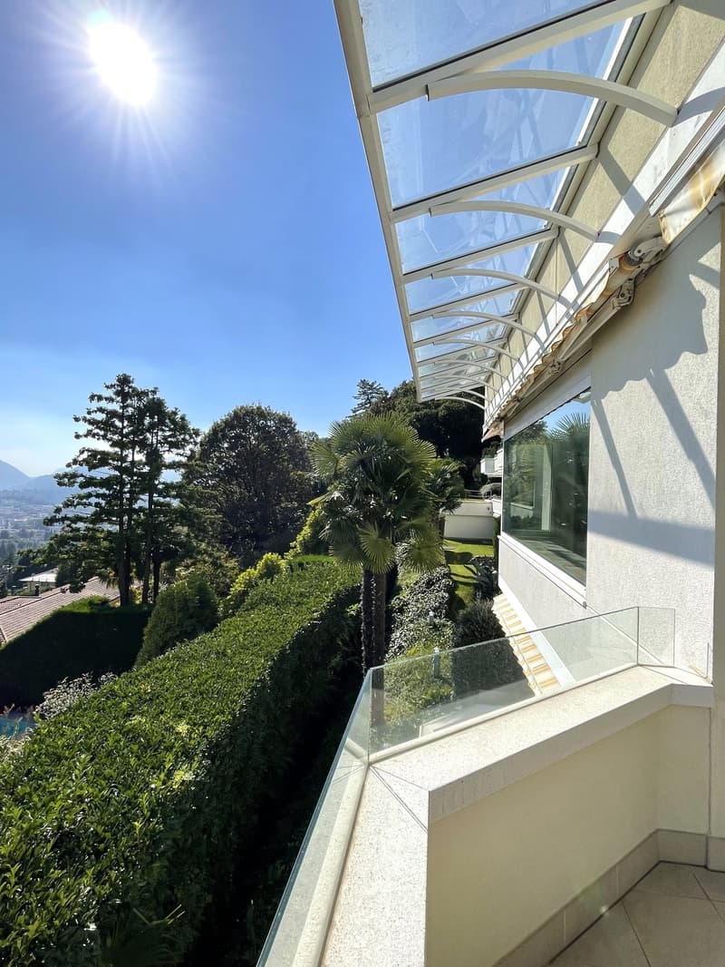 A balcony with a glass railing and a glass canopy, offering a view of a lush garden and distant mountains.