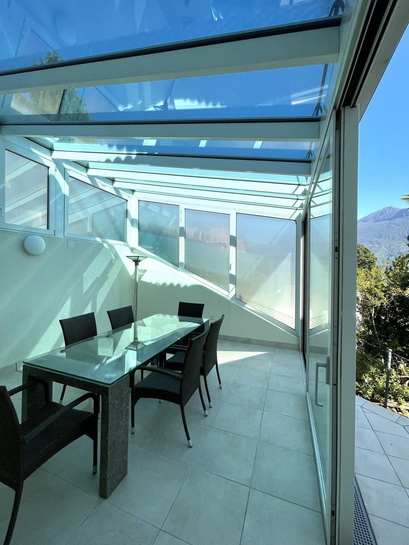 Glass-walled terrace with a dining table and chairs, a floor lamp, and a glass door leading to the outside.