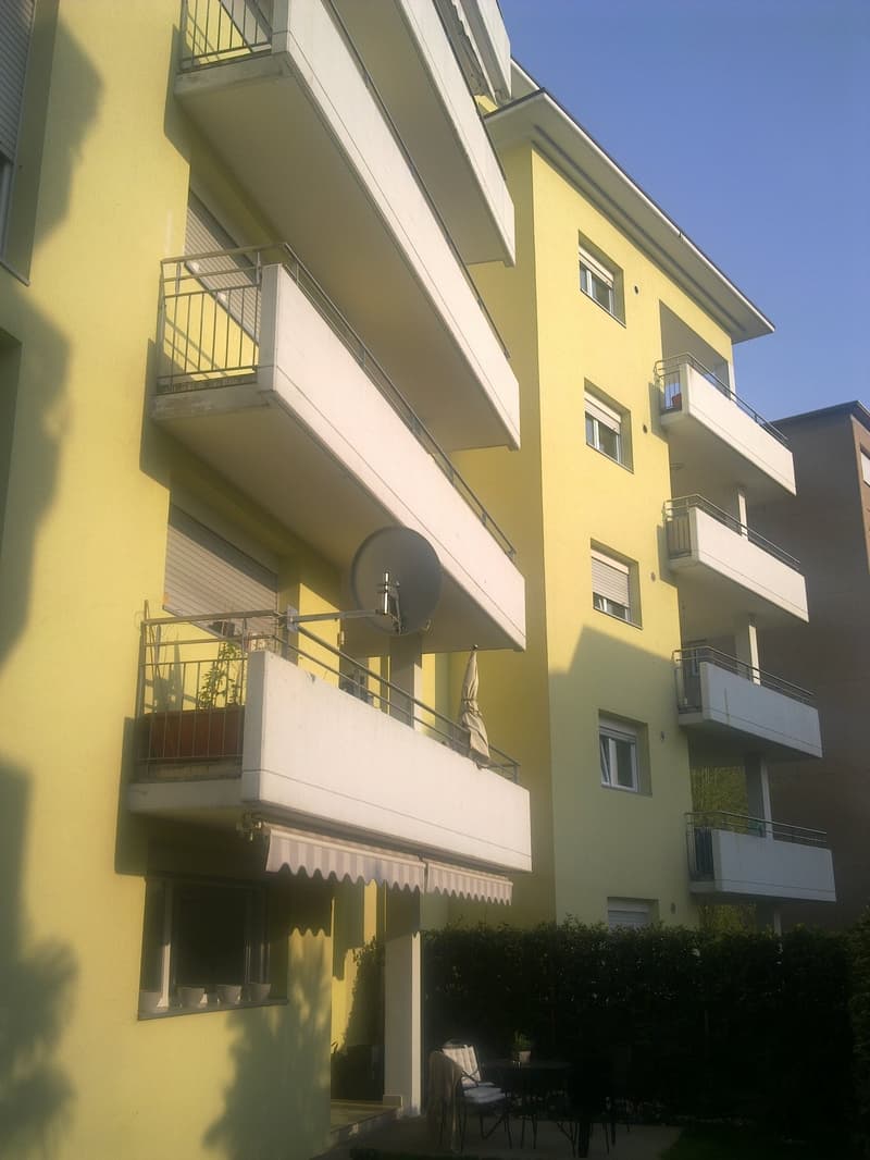 Yellow building with balconies, white awnings, glass doors, metal railings, metal satellite dish, garden with plants, outdoor table, chairs, and plants