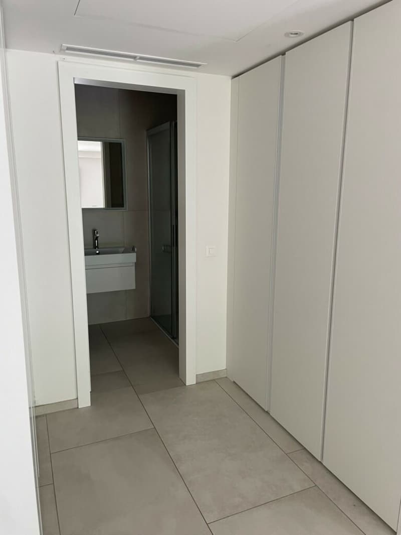 Empty bathroom with gray tiled floor, white walls, modern fixtures, glass-enclosed shower, mirror, and sink.