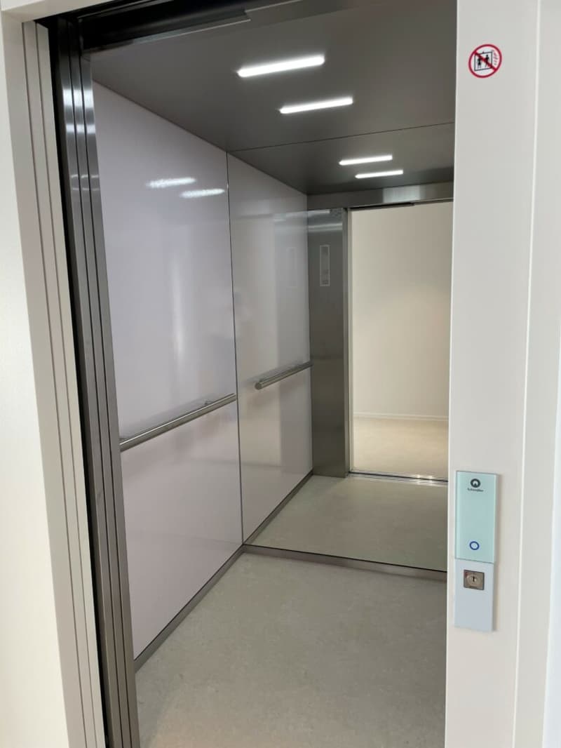 The image shows an elevator with a steel frame, interior illuminated by ceiling lights, and a digital control panel on the outside.