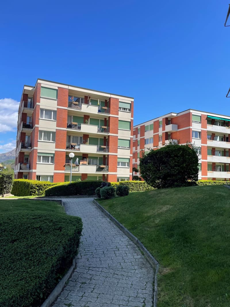 Multi-story apartment building with brick and stucco exterior, balconies on each unit, surrounded by landscaped grounds with a paved walkway.