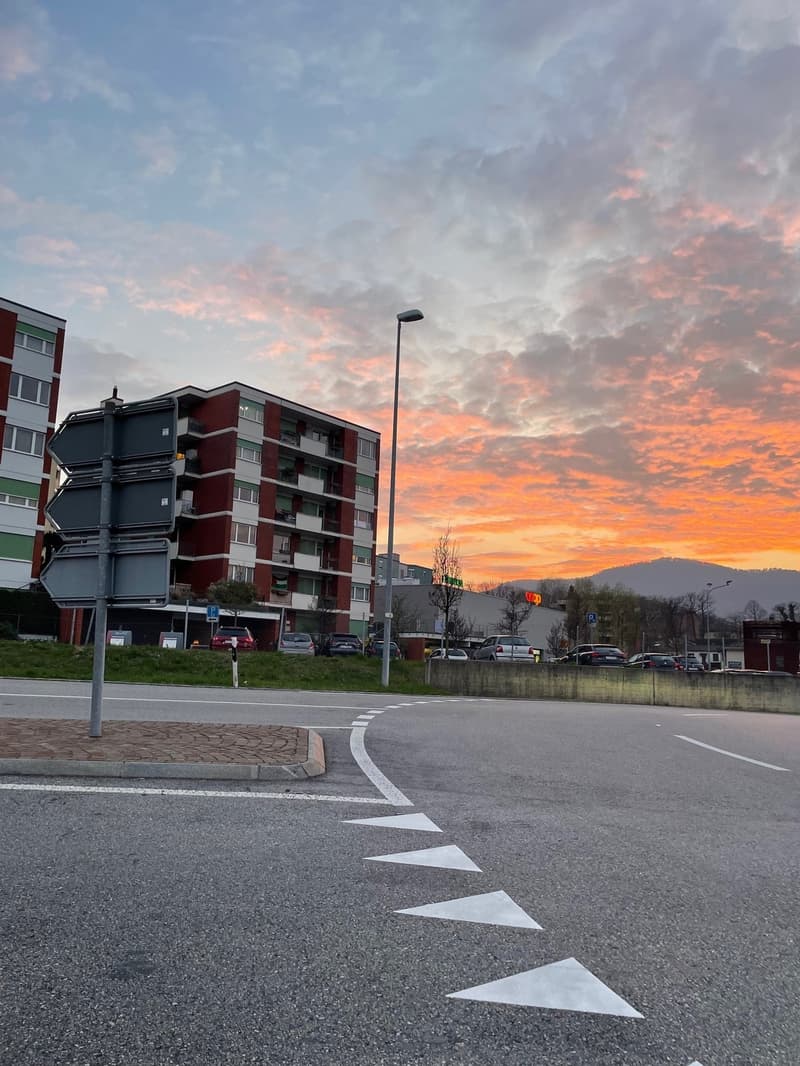 The image shows a residential area with multi-story apartment buildings, a road with a crosswalk, and a dramatic sunset sky with colorful clouds.