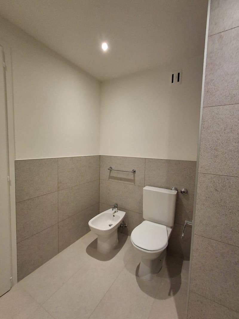 modern minimalist bathroom with white ceramic tile flooring, wall-mounted bidet and toilet, small shower enclosure, gray tiled walls, white ceiling, and light.